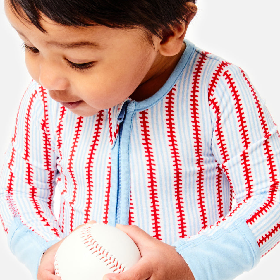 A toddler wearing a light blue and white zip-up pajama shirt with red baseball-stitch print details, holding a baseball against a clean white background. The Clover Baby & Kids logo is visible in the bottom-left corner.
